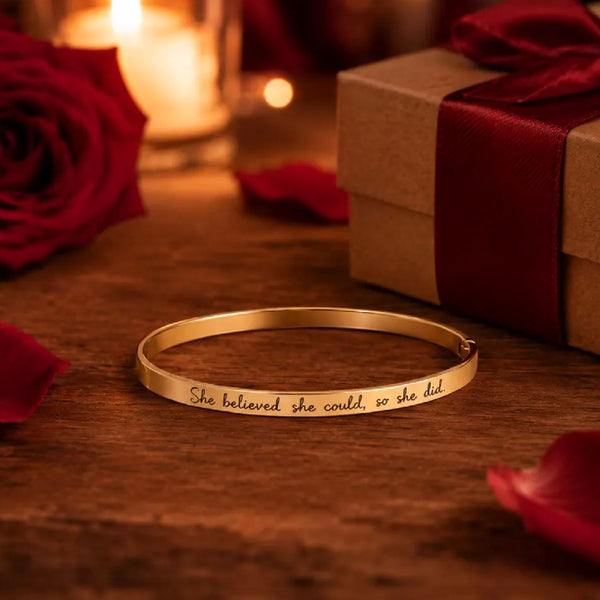 A gold-toned metal bangle bracelet engraved with the phrase 'She believed she could, so she did' in black cursive script, resting on a wooden surface. The scene is styled with red rose petals, a red rose, a brown gift box with a red ribbon, and a warm glowing candle in the background.