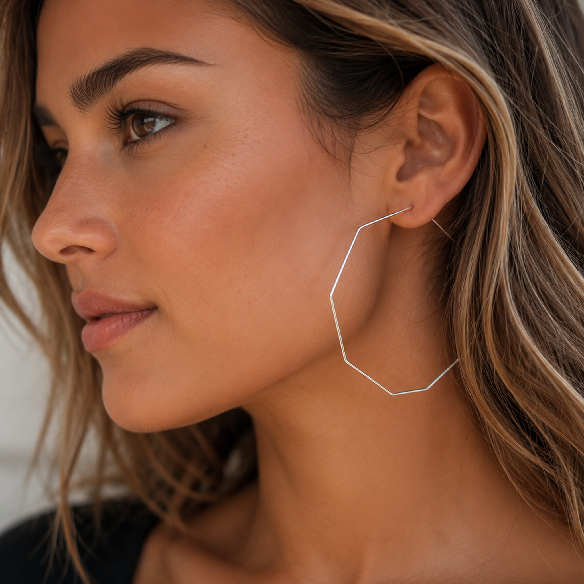 A close-up side profile of a woman wearing a delicate, thin silver geometric hoop earring that follows the curve of her ear.