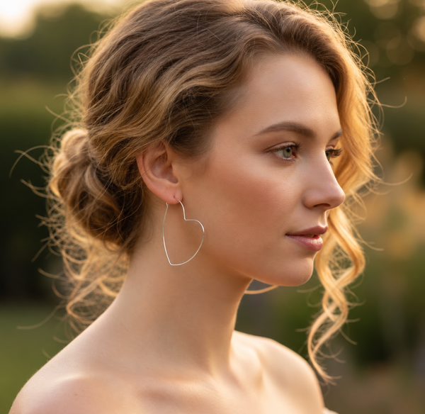A woman in profile wearing a delicate silver heart-shaped hoop earring, photographed outdoors with a soft, blurred green and warm golden background.