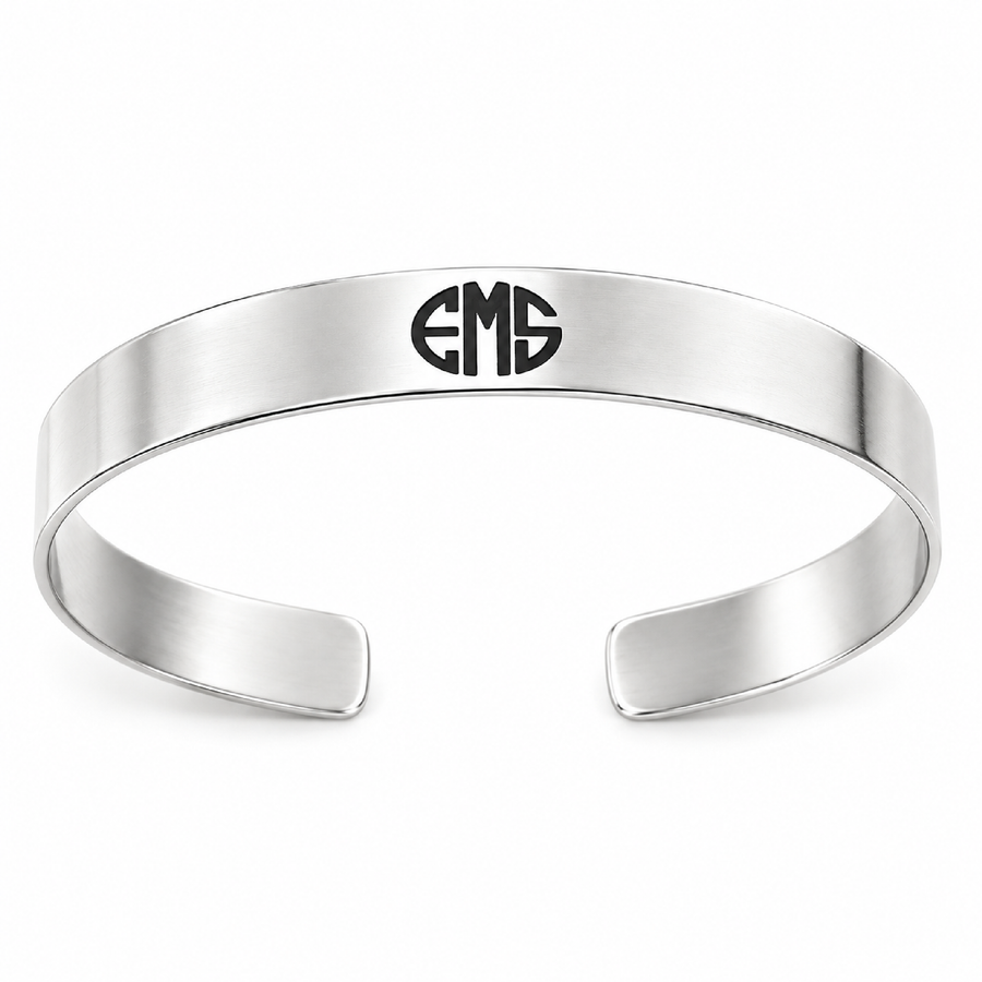 A polished silver cuff bracelet with a black monogram engraving, presented inside a gray gift box with a red ribbon.