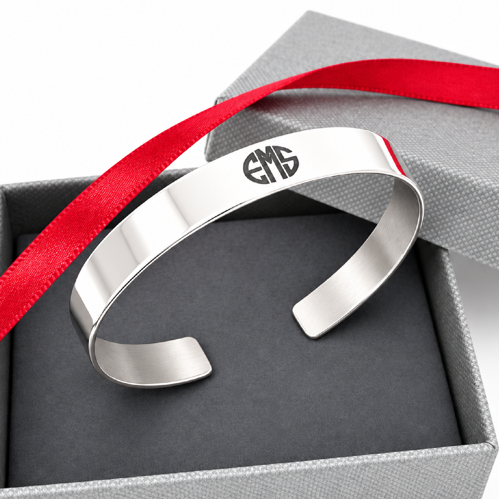A polished silver cuff bracelet with a black monogram engraving, presented inside a gray gift box with a red ribbon.