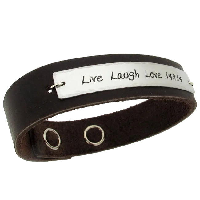 A black leather bracelet with a custom engraved message 'Live Laugh Love 1981' on a white metal plate, featuring two metal eyelets for adjustability.
